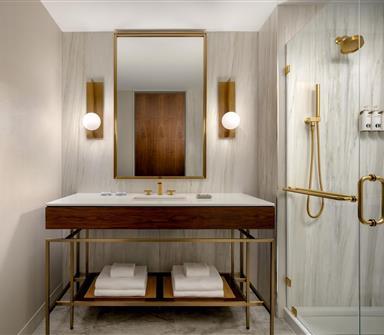 Bright hotel bathroom with wood vanity, white countertop, large brass-framed mirror, globe sconces, and glass shower with brass fixtures.