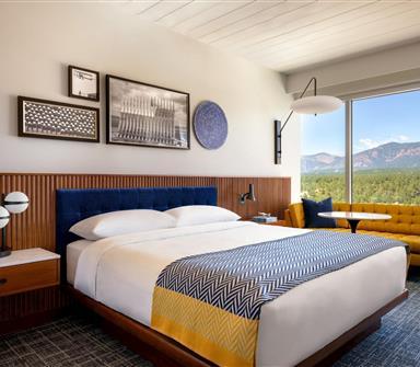 King guest room with white bedding, blue-and-yellow patterned throw, blue headboard, warm wood paneling, mustard sofa, and expansive mountain view.