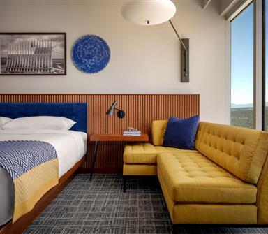Guest room seating area with mustard-yellow sectional sofa, navy pillow, wood-paneled wall, blue headboard, and large window overlooking mountains.