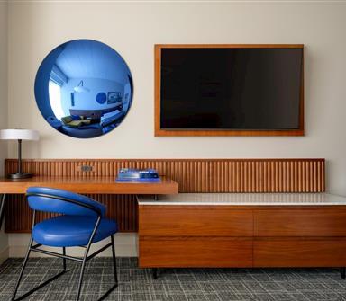 Guest room workspace with blue upholstered chair, floating wood desk, round blue-tinted mirror, wall-mounted TV, and low wood dresser.