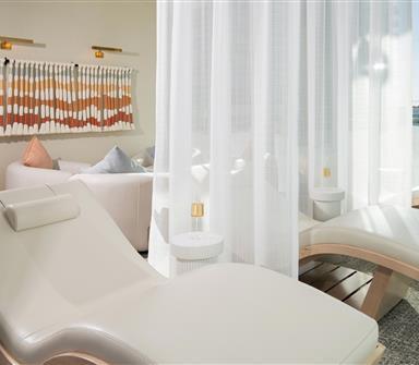 Sunlit spa relaxation room with white contoured lounge chairs, sheer privacy curtains, small side tables, and colorful woven wall art.