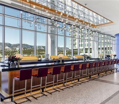 Long contemporary bar with burgundy stools, wood and brass finishes, suspended glass lighting, and floor-to-ceiling windows overlooking the mountains.