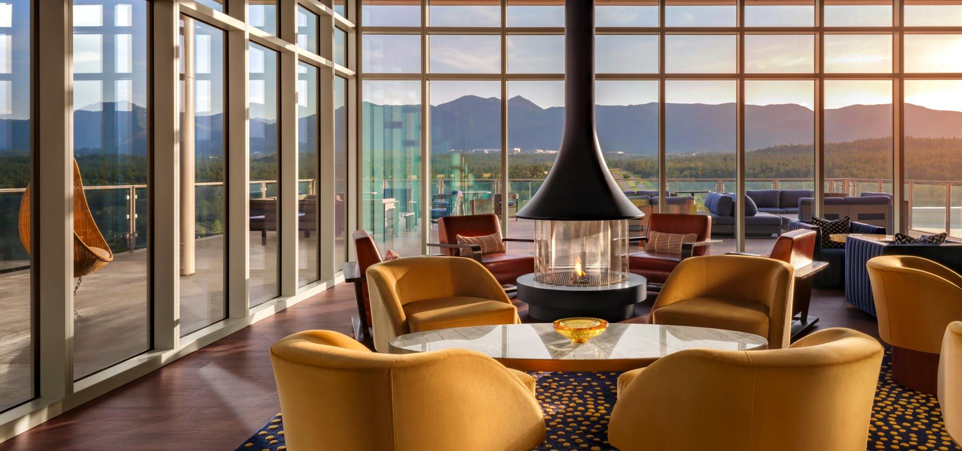A modern lobby lounge with a fireplace, yellow chairs, and floor-to-ceiling windows showing a mountain view.