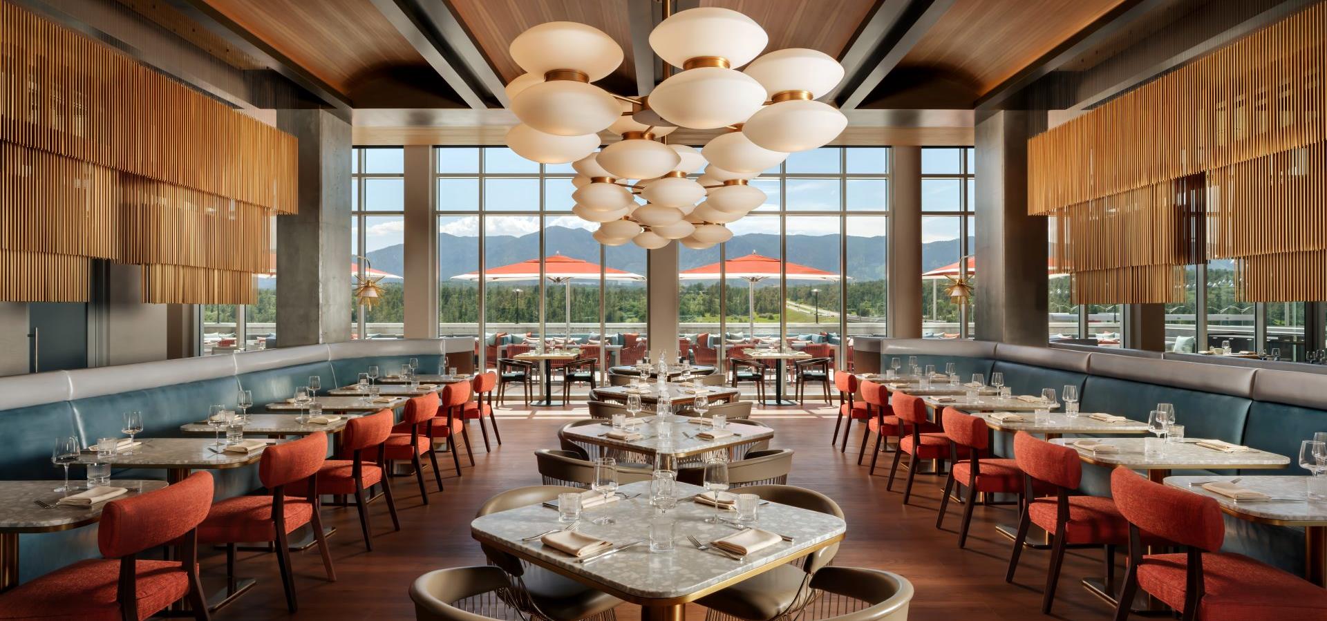 A dining room with a large window showing a mountain view, round tables with chairs, and a unique lighting fixture.