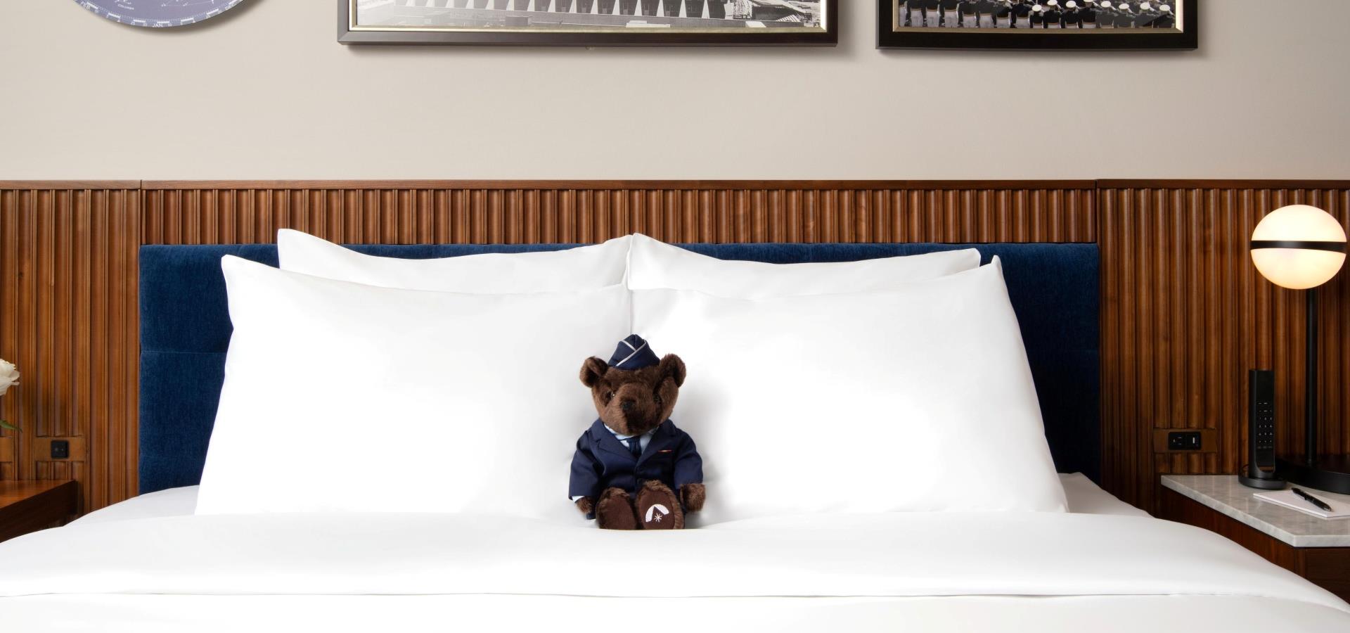 A neatly made bed with a stuffed bear sitting on it, in a room with wood paneling and modern lighting.