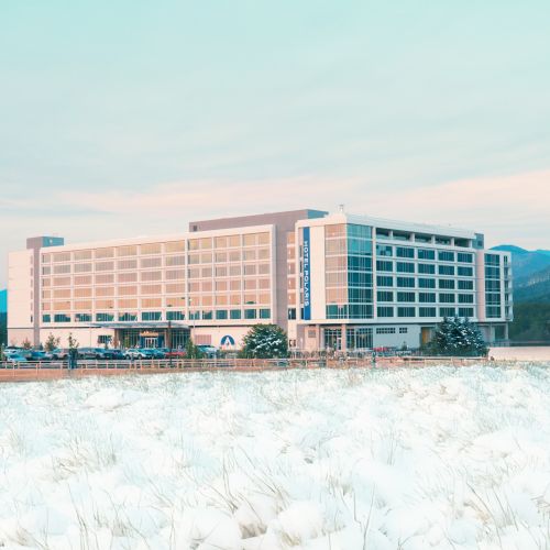 A large, modern hotel sits by a snowy field with mountains in the background, under a pale blue sky.