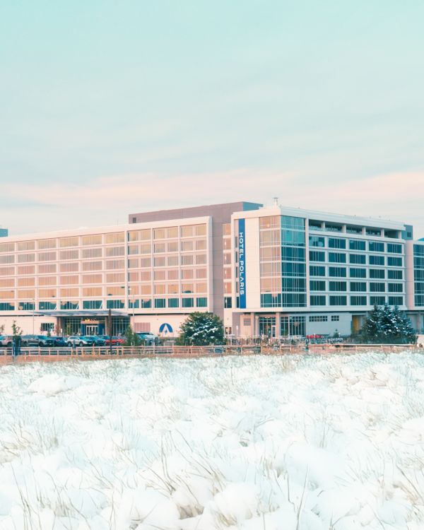 A large, modern hotel sits by a snowy field with mountains in the background, under a pale blue sky.